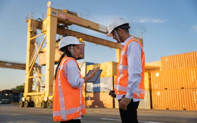 Zwei Personen in Sicherheitskleidung sprechen miteinander vor einem Containerterminal.