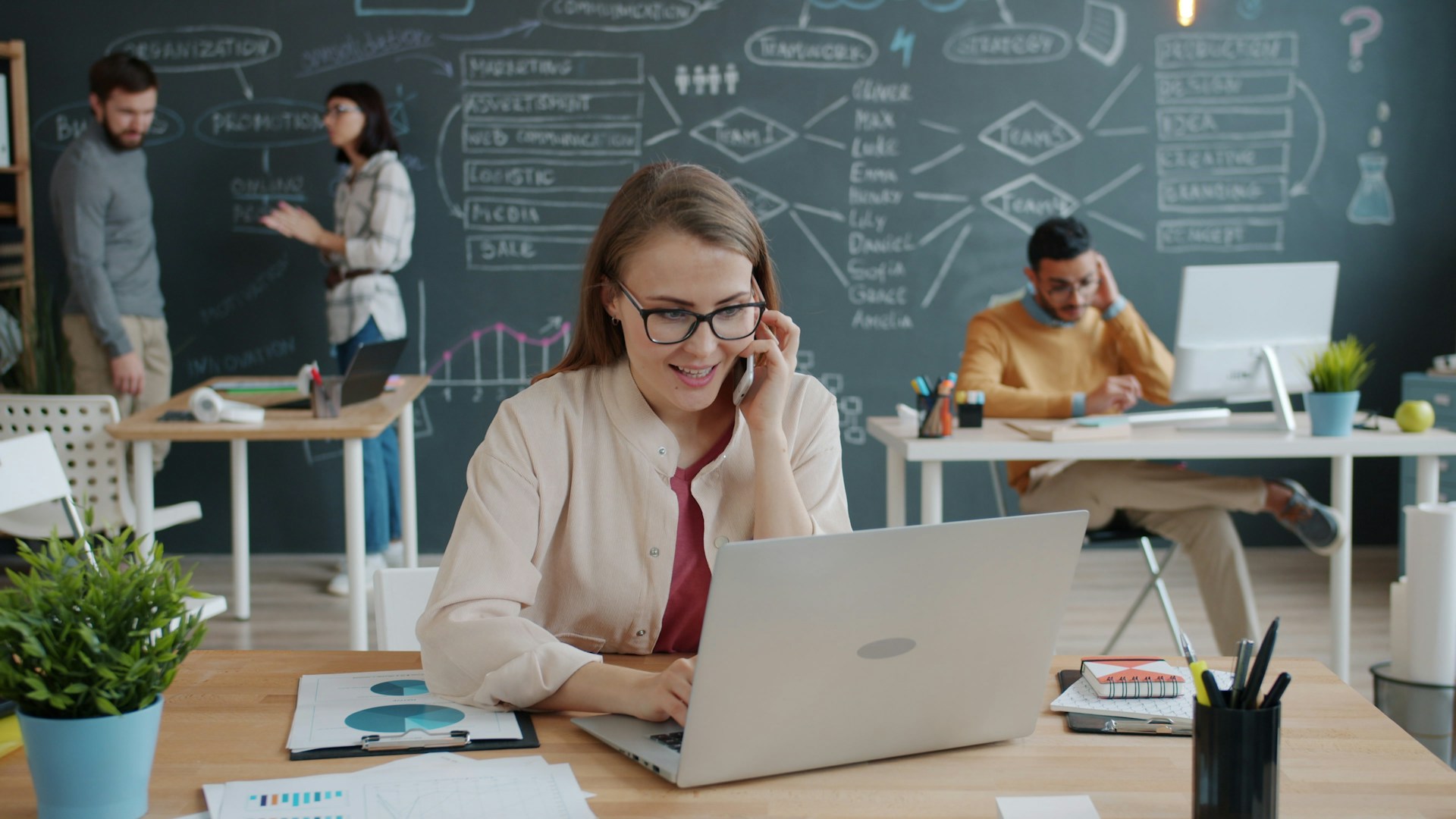 Vier Mitarbeiter in einem Büro. Zwei sitzen an einem Schreibtisch und arbeiten an ihren Computern. Im Hintergrund unterhalten sich die anderen beiden.